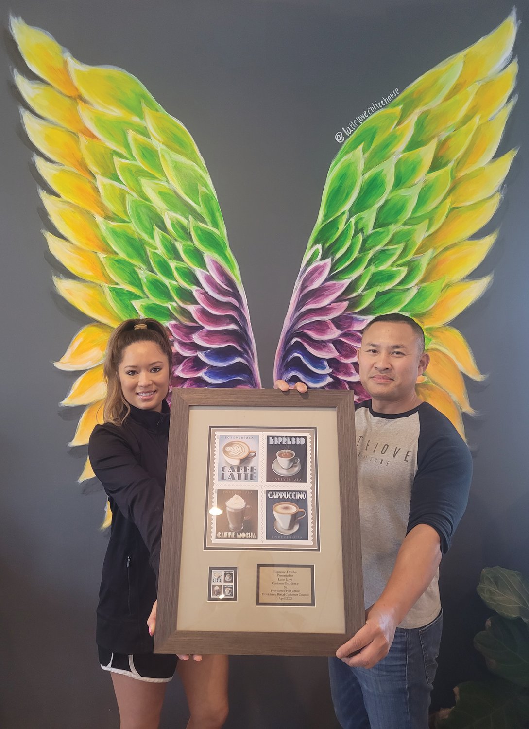 COFFEE ANGELS: Tony and Elizabeth Phouaykoumpha stand against one wall in their Johnston coffee shop. A pair of brilliantly colored, painted angel wings frame the Scituate couple as they hold a framed bit of appreciation from the Providence Postal Customer Council. Their first (of two) location opened in 2018 in Cranston. (Sun Rise photo by Rory Schuler)